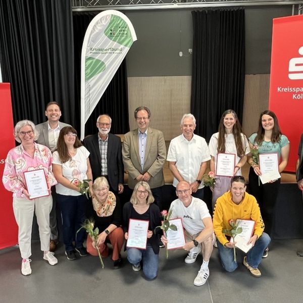 Gruppenfoto von etwa 13 Personen bei einer Ehrungsveranstaltung des Kreissportbundes. Mehrere Geehrte halten Urkunden und Rosen in den Händen. Im Hintergrund sind zwei rote Roll-ups der Kreissparkasse Köln sowie eine Beachflag des Kreissportbundes zu sehen. Die Gruppe ist in zwei Reihen angeordnet – stehend und kniend – in einem Veranstaltungsraum.