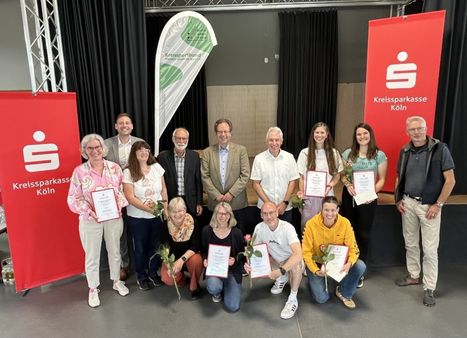 Gruppenfoto von etwa 13 Personen bei einer Ehrungsveranstaltung des Kreissportbundes. Mehrere Geehrte halten Urkunden und Rosen in den Händen. Im Hintergrund sind zwei rote Roll-ups der Kreissparkasse Köln sowie eine Beachflag des Kreissportbundes zu sehen. Die Gruppe ist in zwei Reihen angeordnet – stehend und kniend – in einem Veranstaltungsraum.